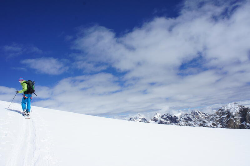 Sur une pente neigeuse, un skieur prend la pause au soleil. Au fond on aperçoit les montagnes rocheuses enneigées. Le ciel passe d’un bleu vif à des nuages.